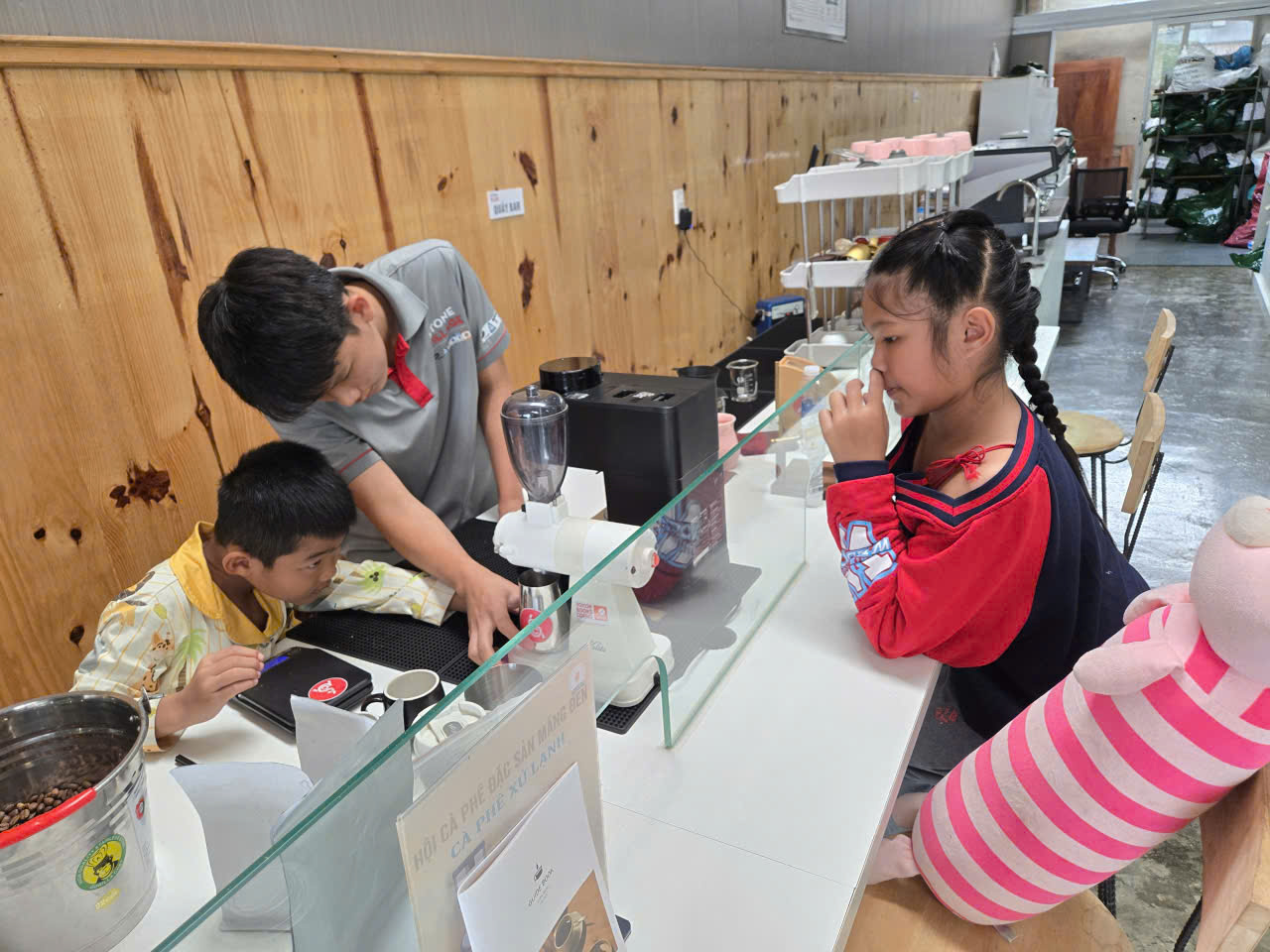 Giảng viên hướng dẫn các bé barista nhí tại quầy bar, một bé quan sát máy xay, một bé ngửi hương cà phê.