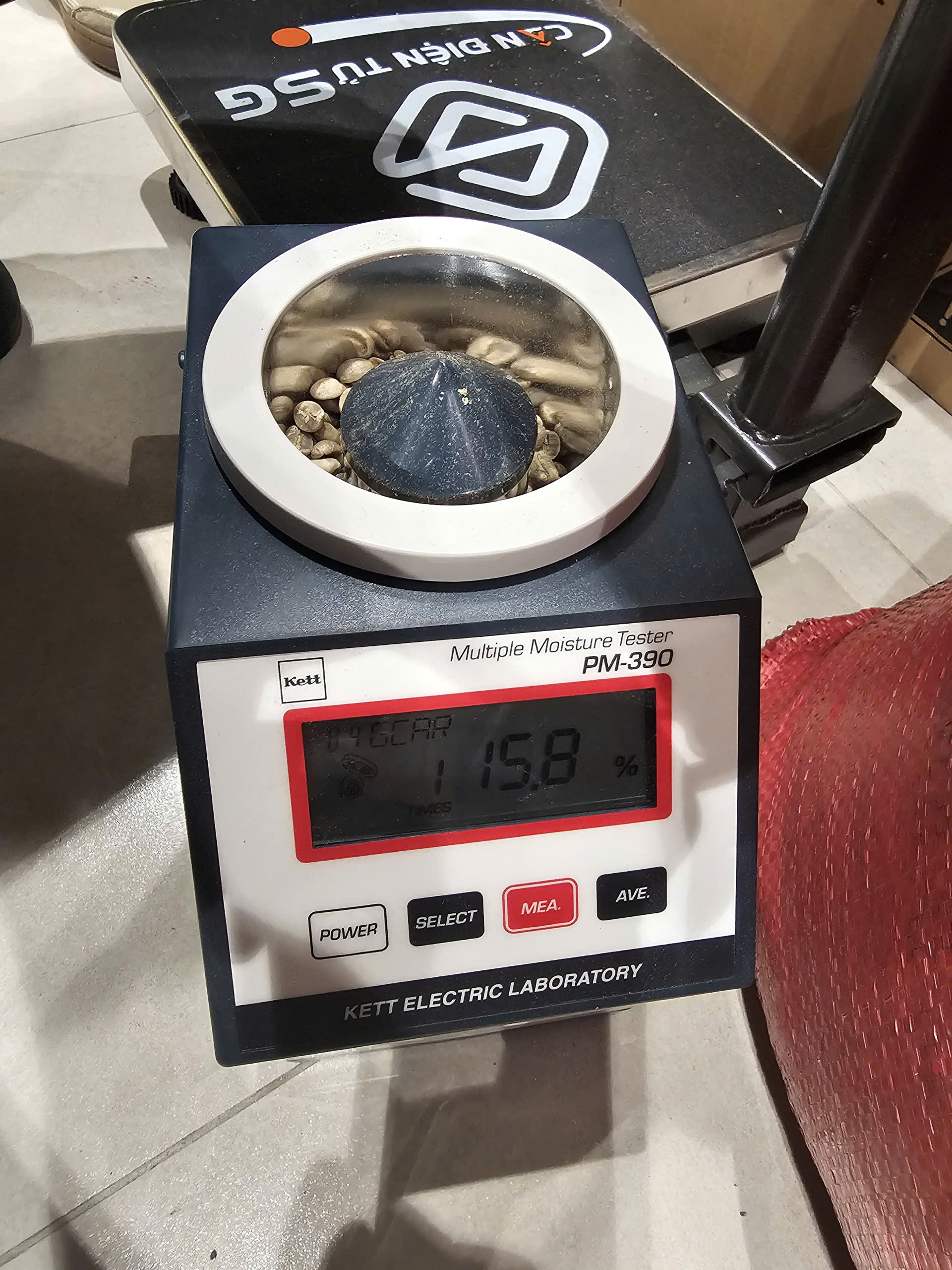Máy đo ẩm Kett PM-390 đặt cạnh cân điện tử và bao cà phê khi kiểm tra ẩm độ nhân xanh nhập kho