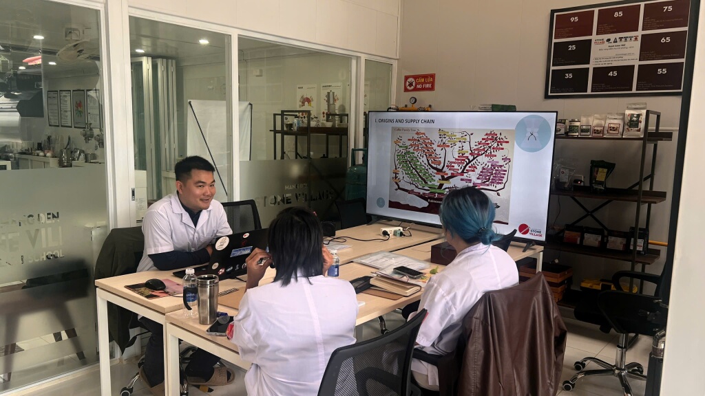 Buổi lý thuyết trong lớp Sensory tại Lab Măng Đen, màn hình chiếu phần Origins and Supply Chain kèm sơ đồ cây gia phả cà phê. Trên tường là bảng thang màu rang từ 25 tới 95.