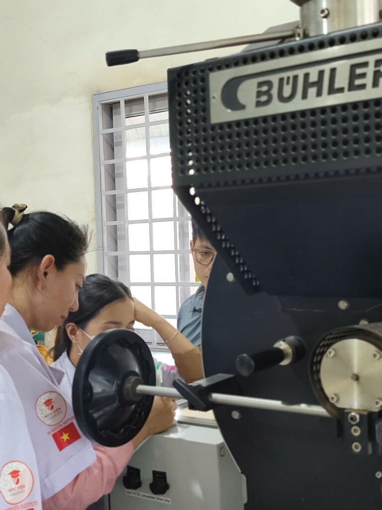 Nhóm kỹ thuật và học viên cùng quan sát cửa lấy mẫu của máy rang Bühler trong buổi hỗ trợ tại xưởng