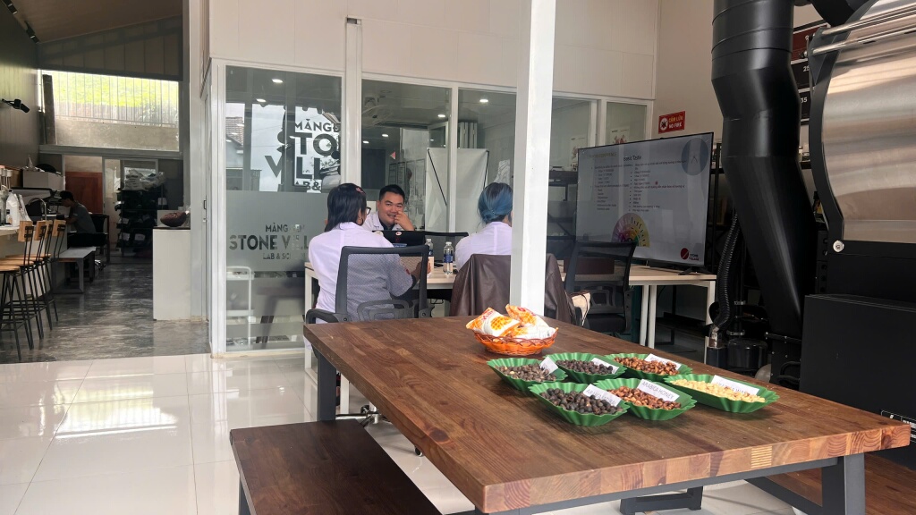 Bàn bày các khay mẫu hạt đặt sẵn trong phòng học, phía sau là máy rang và màn hình bài giảng.