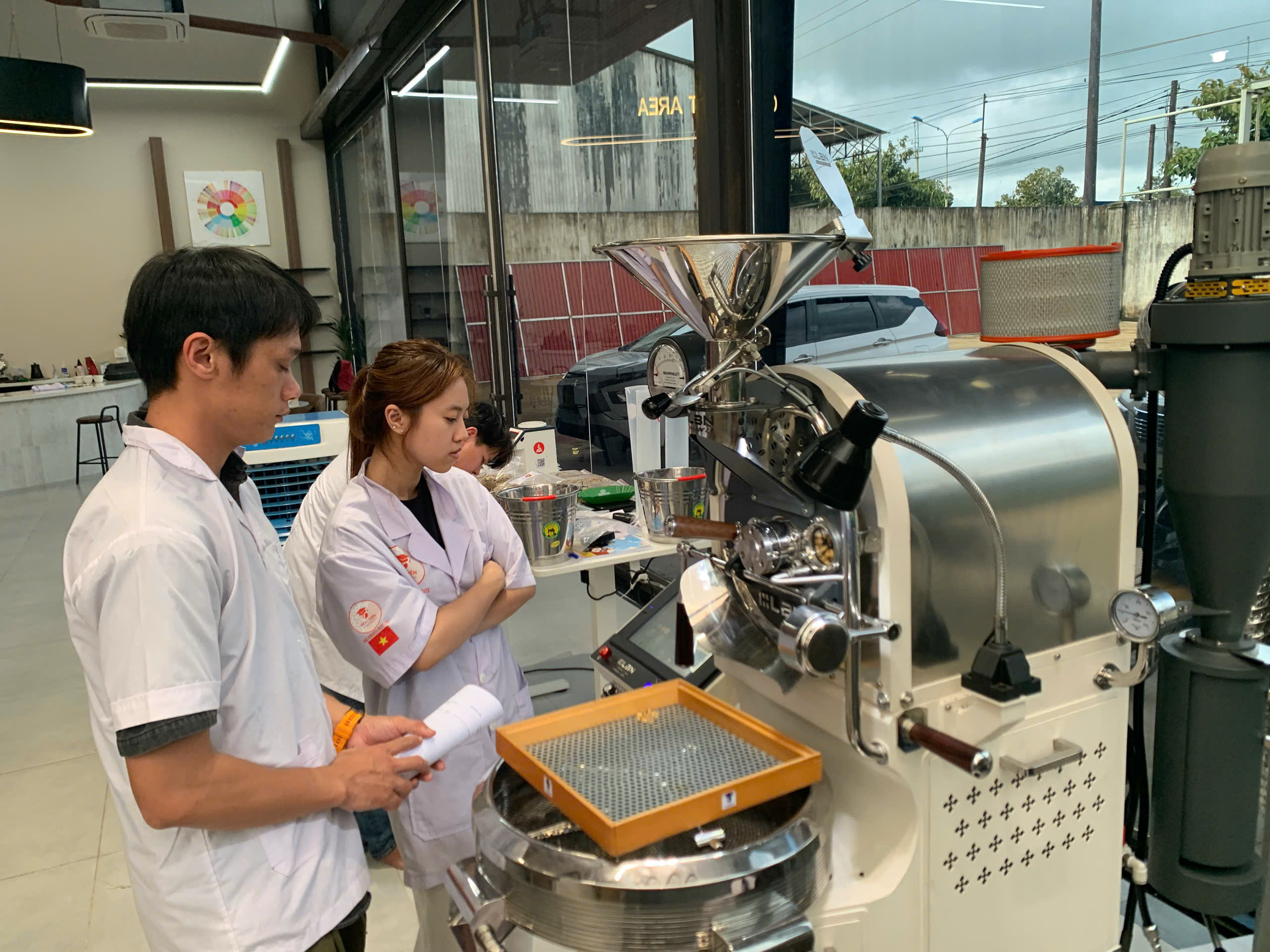Hai học viên cầm khay sàng hạt bên máy rang lớp SCA Roasting Gia Lai