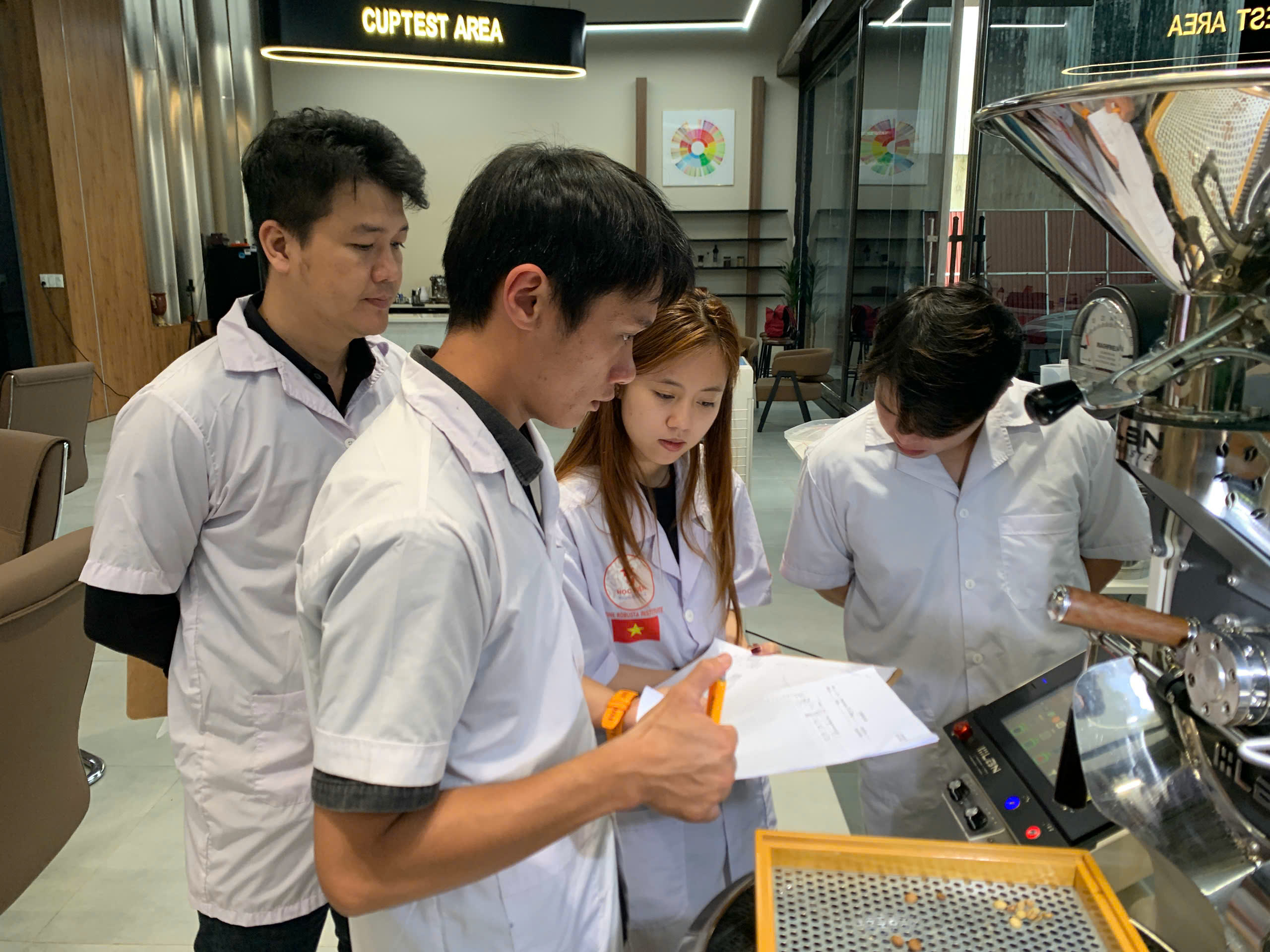 Học viên lớp SCA Roasting Gia Lai đứng bên máy rang phòng lab, cầm phiếu ghi thông số