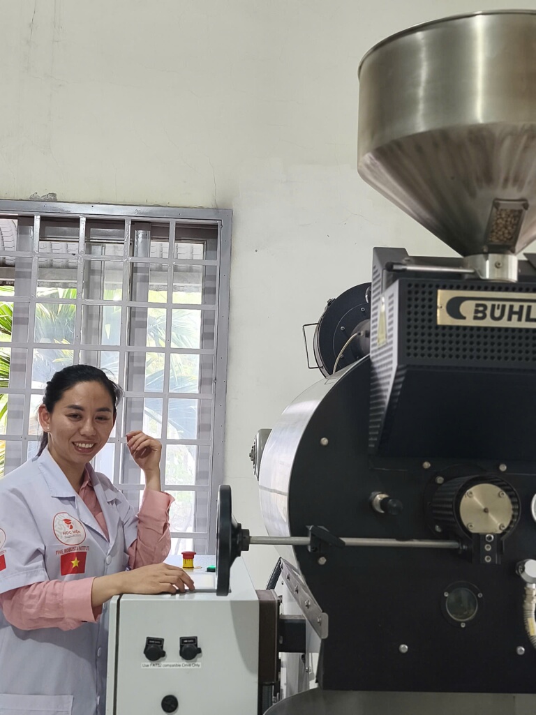 Máy rang cà phê công nghiệp Bühler với phễu nạp nhân xanh phía trên tại xưởng rang của học viên