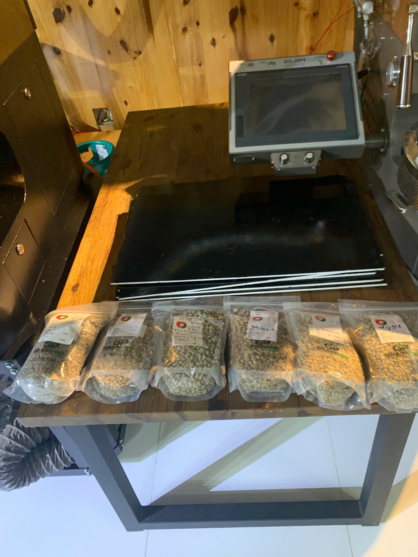 Sáu mẫu nhân xanh đóng túi GrainPro có phiếu ghi thông tin lô, đặt cạnh máy rang mẫu tại Stone Village Coffee Lab